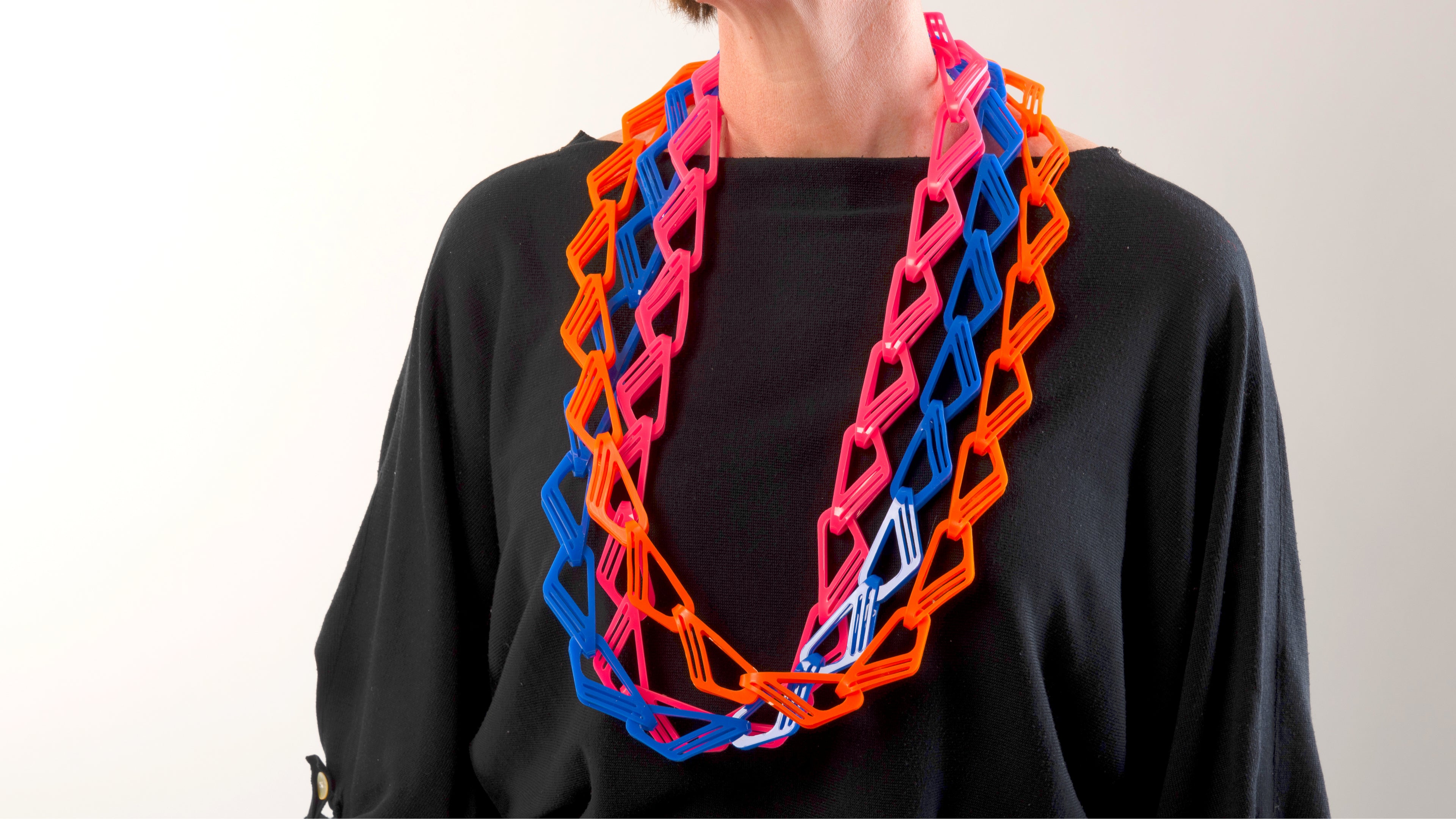 Long and brightly coloured acrylic chain necklaces being worn by a women in a black top. There are three necklaces, one is orange, one is bright pink and one is royal blue.