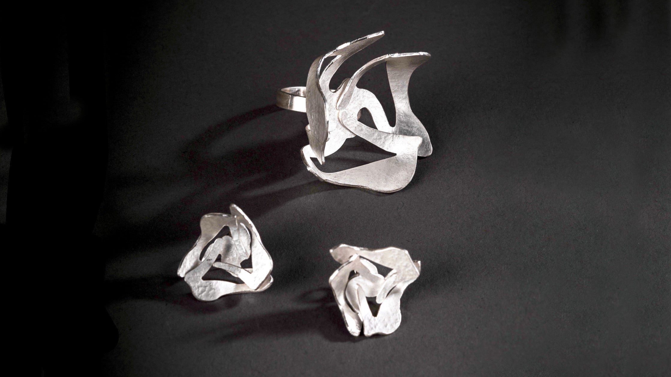 Big silver ring and matching stud style earrings on a black background.  The ring and earrings have a stylised floral shape in textured sterling silver and shiny burnished edges.