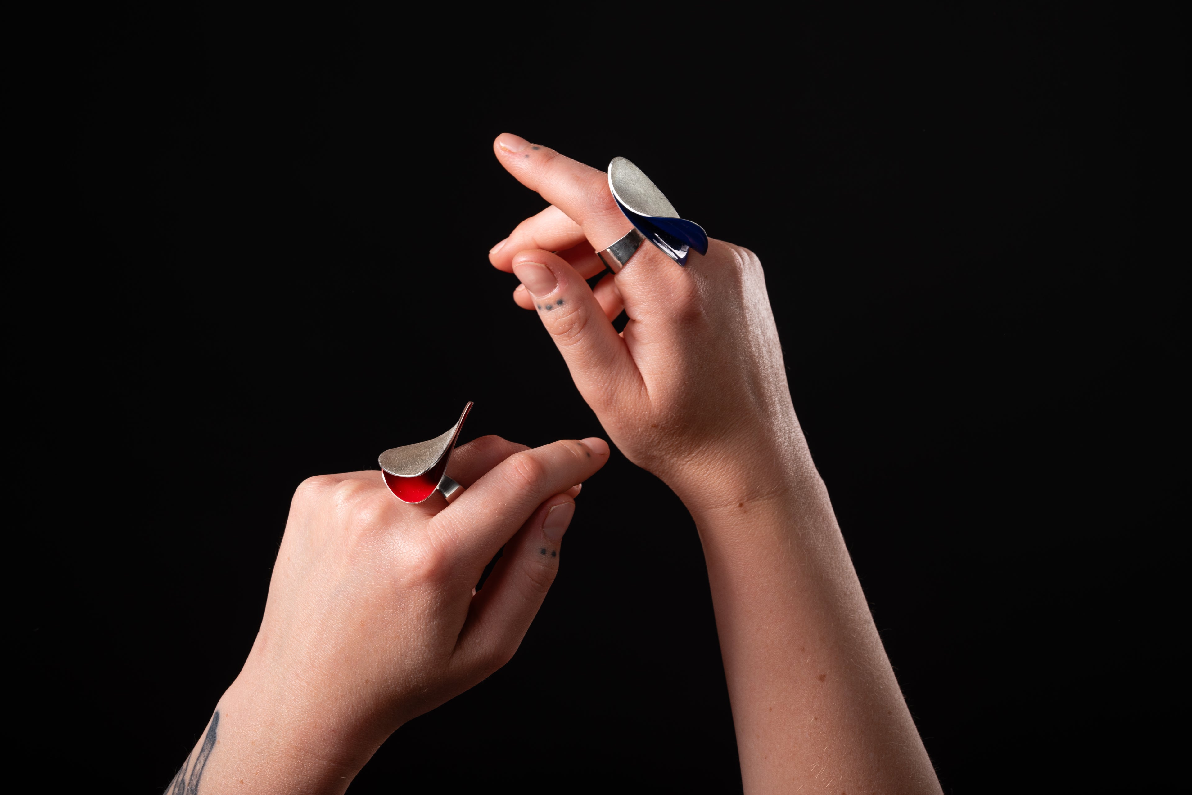 Two large rings being worn on hands which are being held up in the air.  The background is black.  The ring on the left hand is silver with red inside and the ring on the right hand is silver with blue inside.