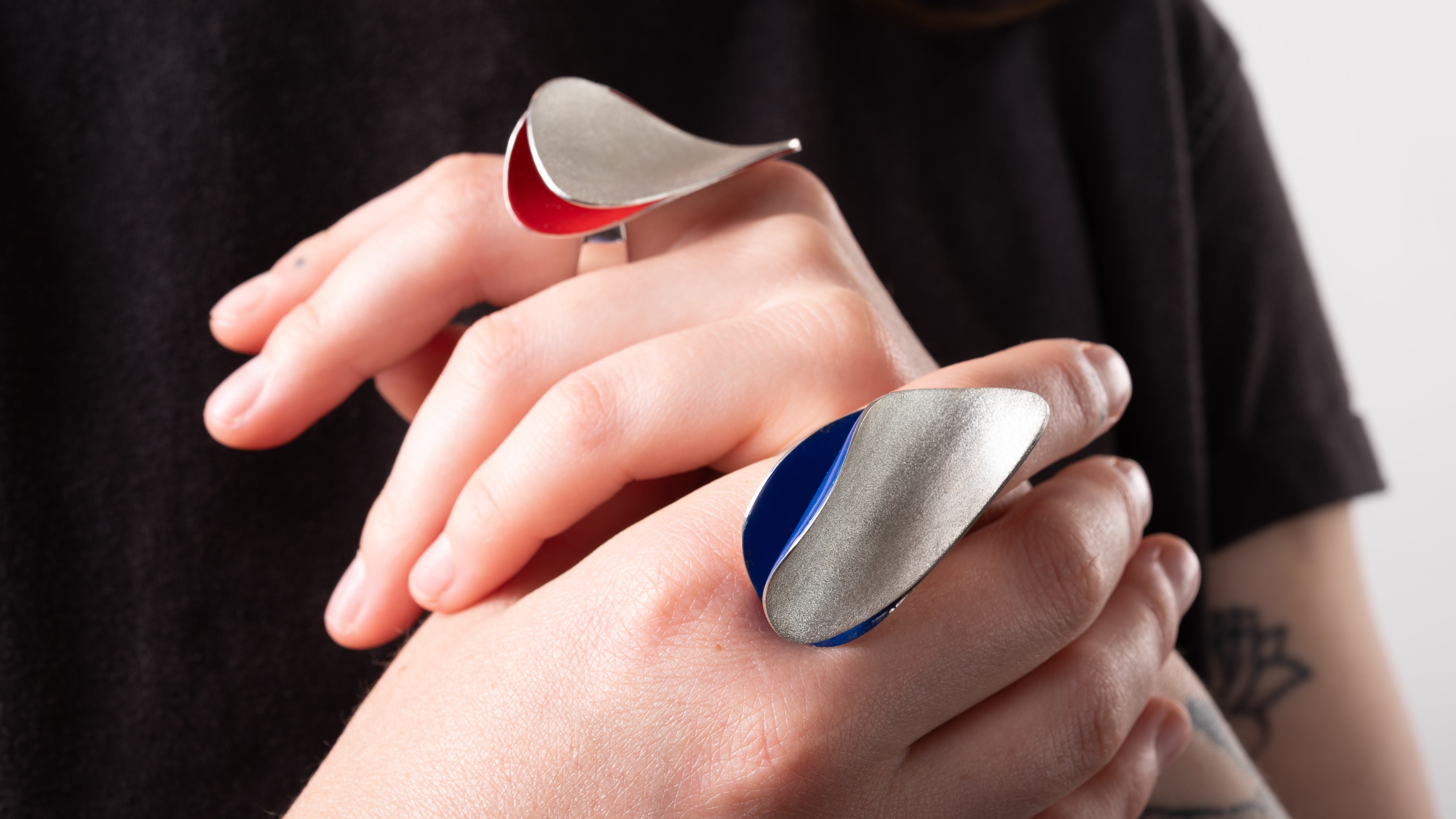 A pair of womens hands crossed in front of her body.  Each hand has one large curved ring.  One ring has a red enameled interior and the other ring has a blue enamelled interior.  The rings are silver with a frosted sparkly finish.