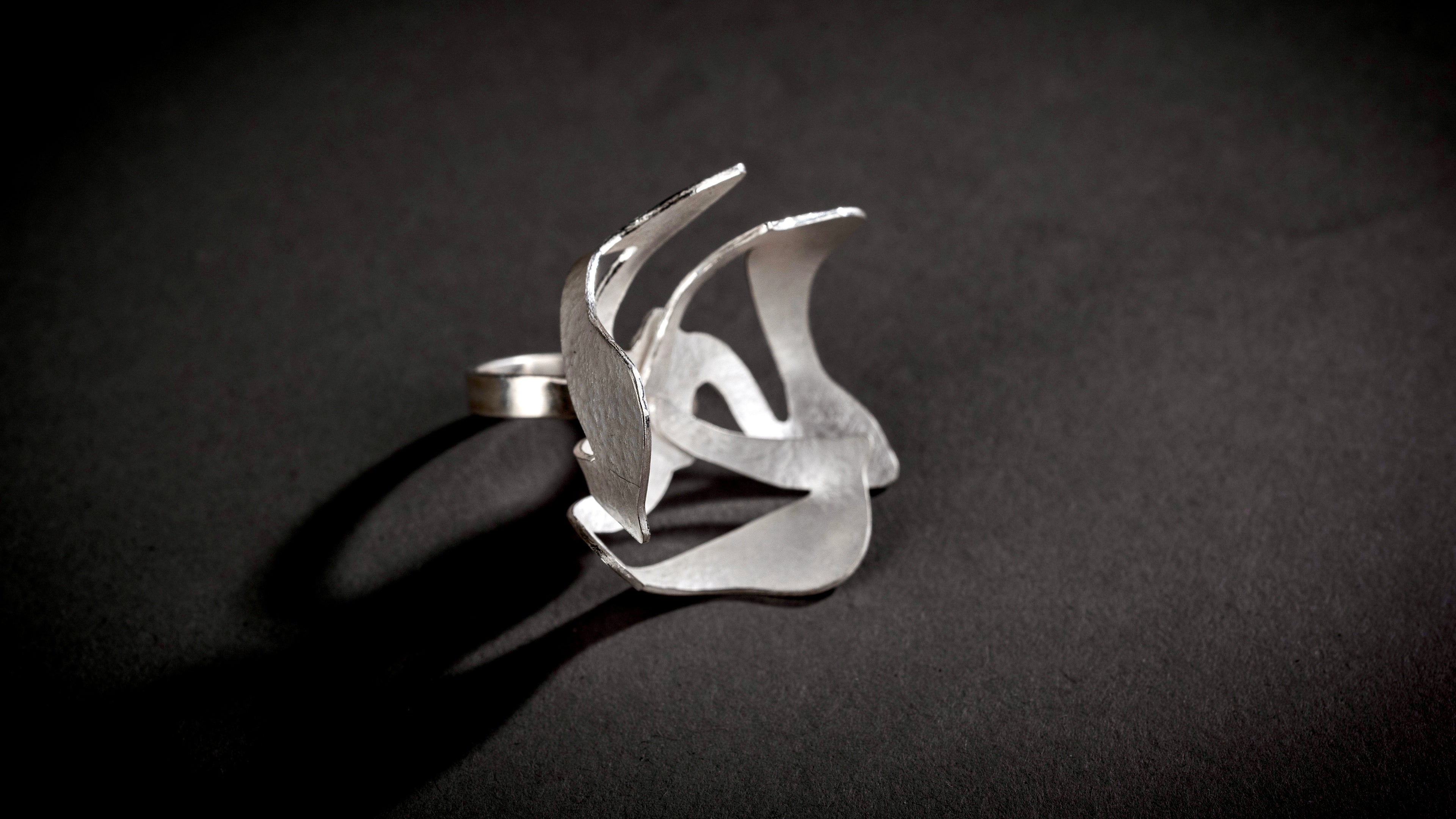 Large silver ring on a black background.  The ring has a large abstract flower shape set on top of a simple silver ring band.  The flower shape has a textured surface and polished shiny edges.
