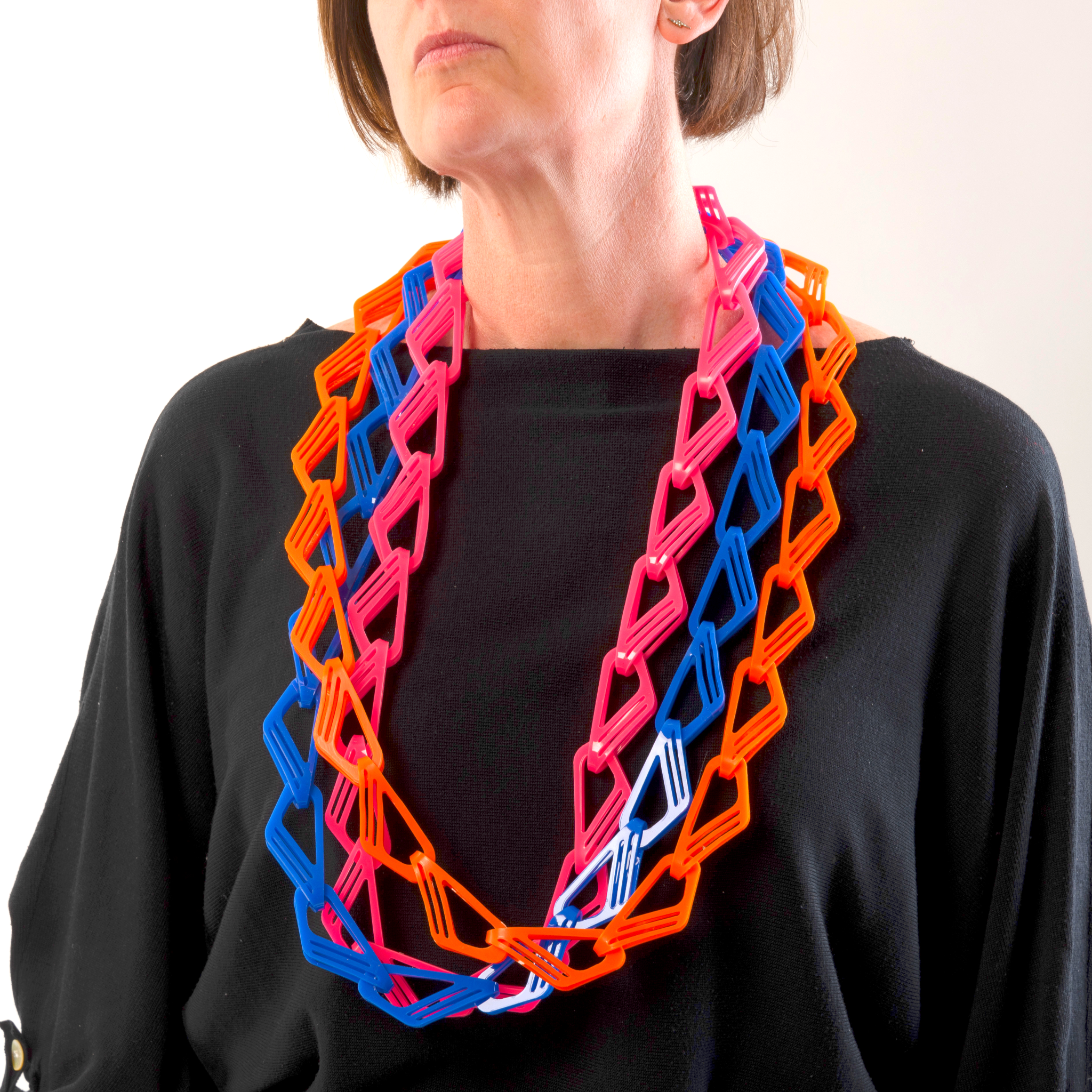 Person wearing a three colorful chain necklaces in brght pink, blue and orange with a plain background.