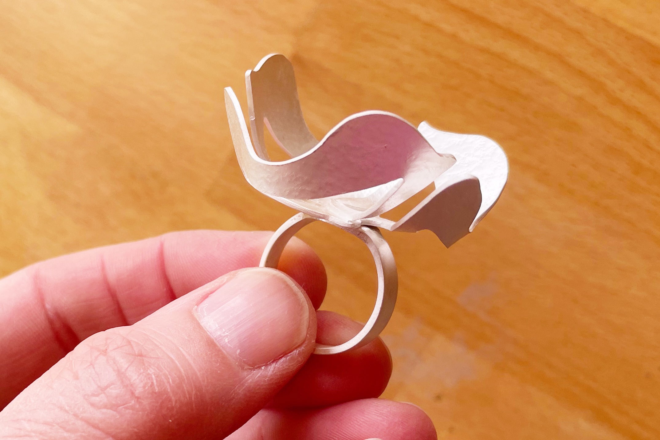 A large silver ring with abstract floral shape atop a silver ring band being held in fingers.