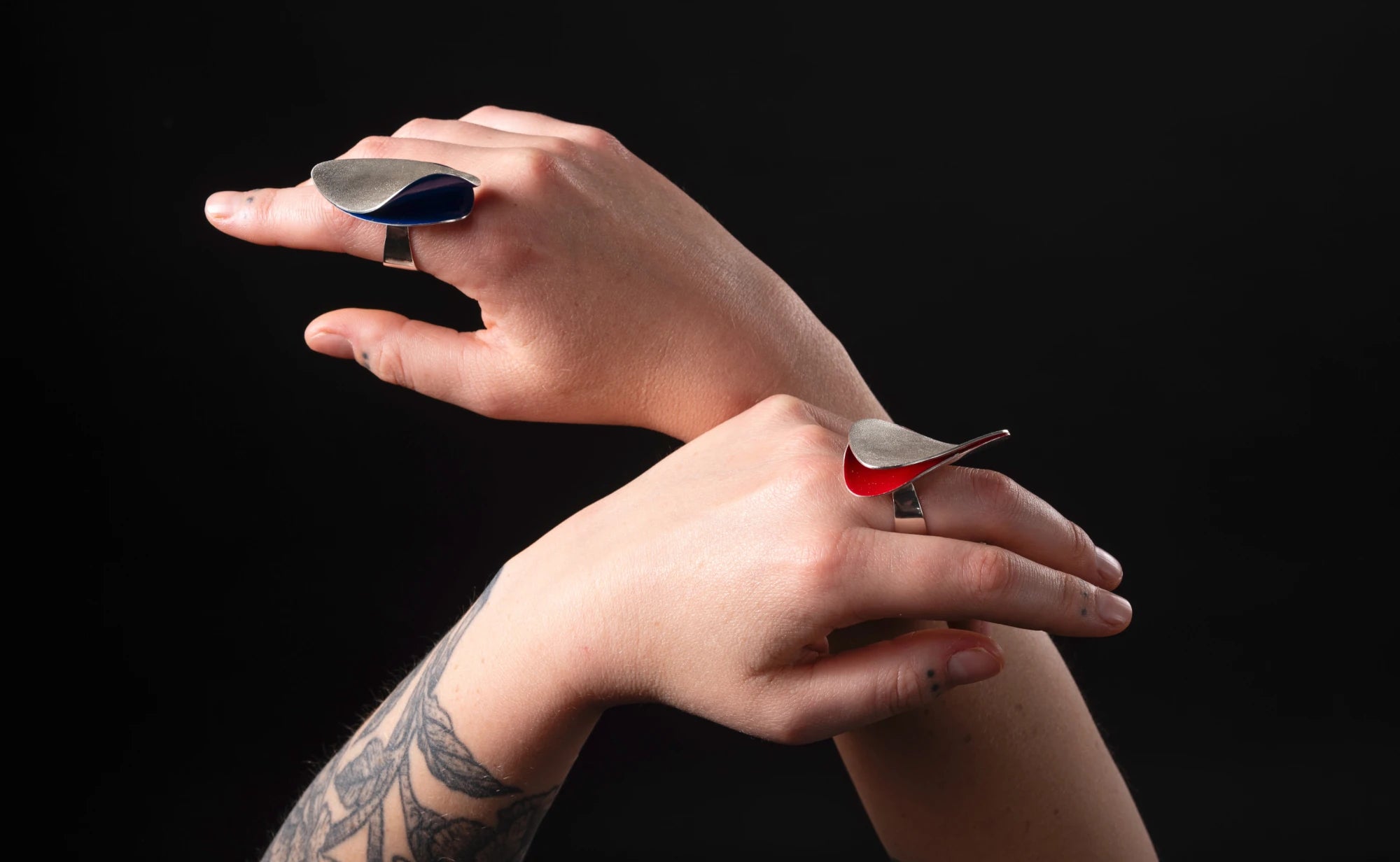 Two hands wearing colorful silver organic scupltural rings on a black background