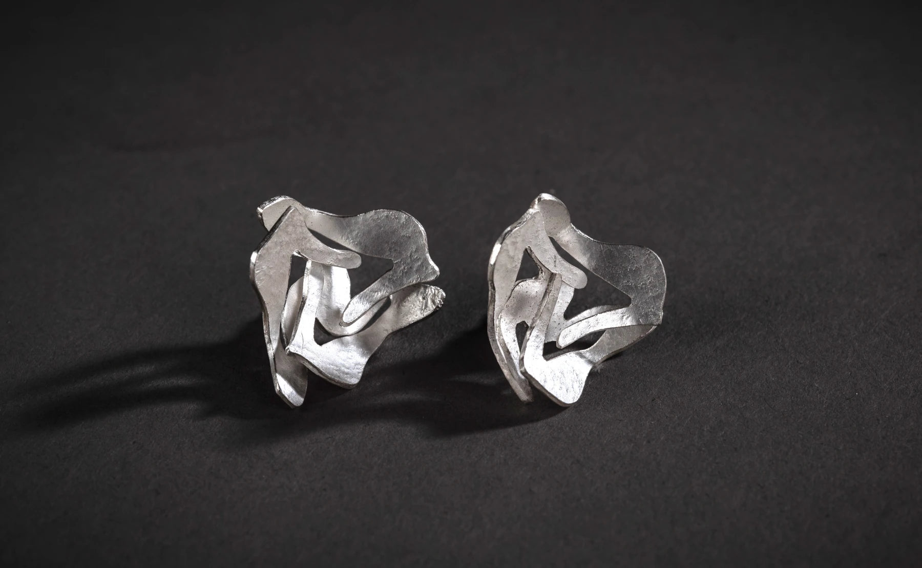 A pair of shiny silver stud earrings with organic shape against a black background.