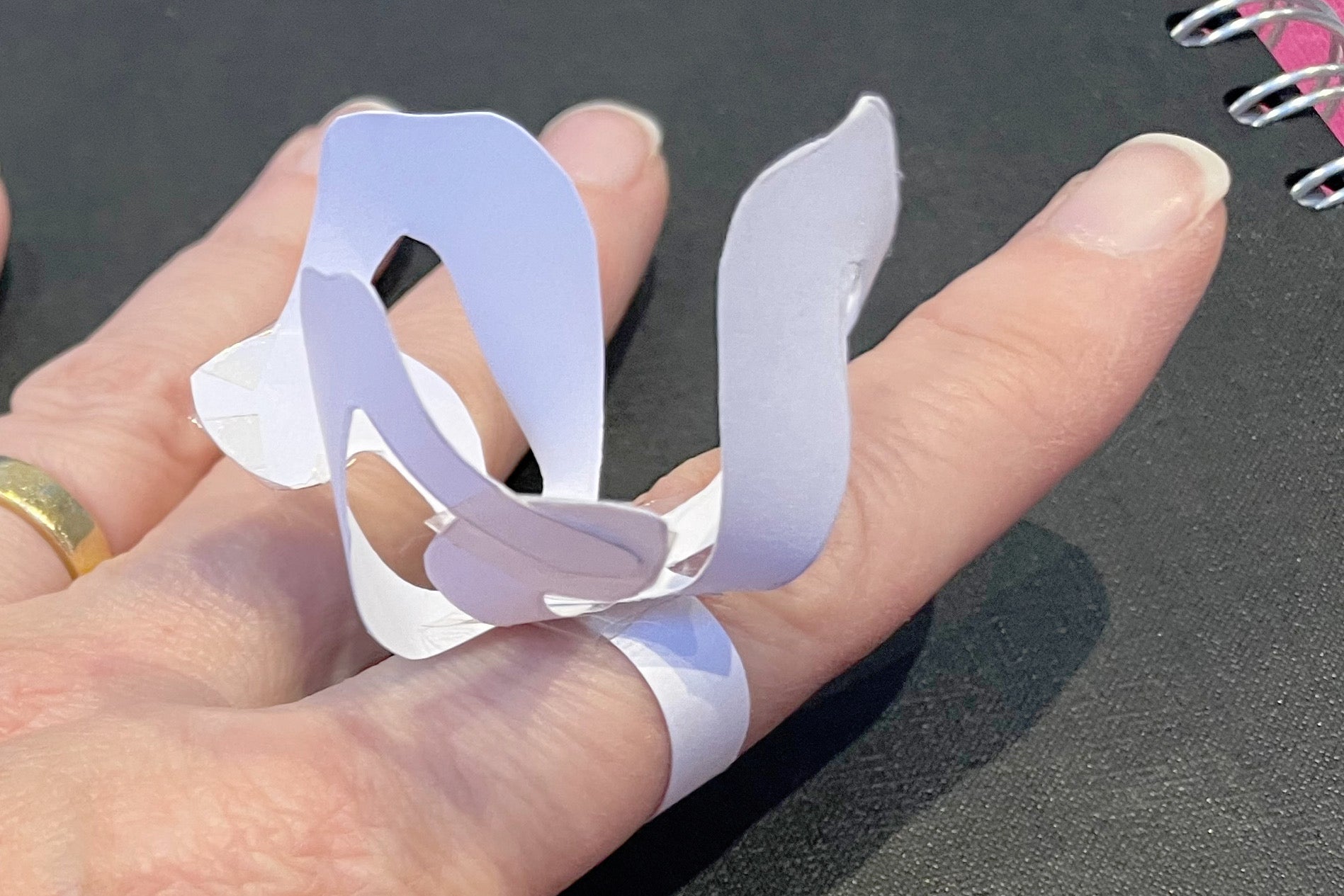 A paper test sample of a large ring with a stylised flower form on a ring band being worn on a hand.
