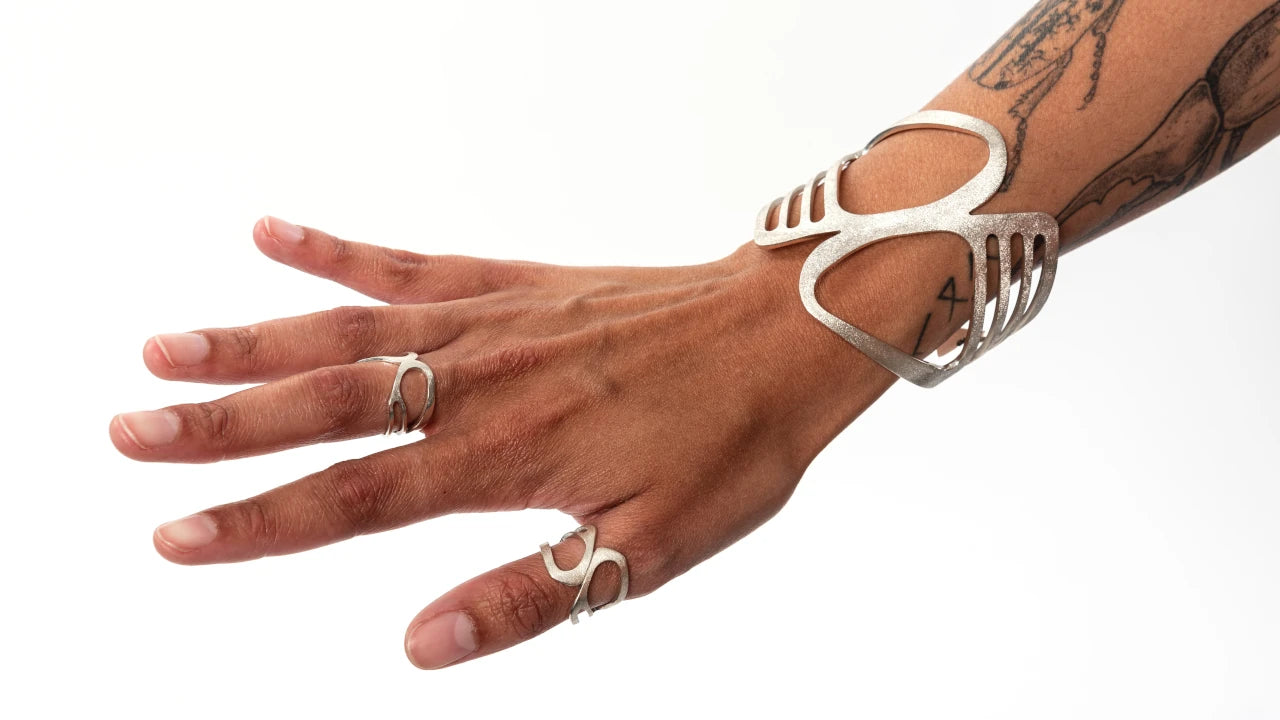 Hand wearing a wide silver bracelet cuff with cut out details, and two organic curved wide rings on a white background