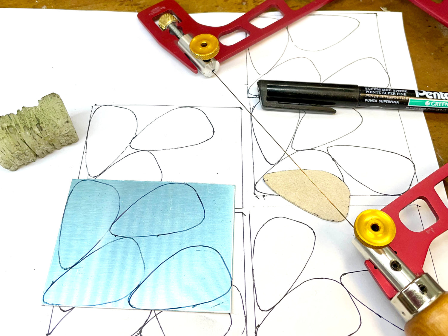 Sterling silver sheet marked up ready to be cut out.  Next to the silver sheet is a jewellers saw, pen and block of beeswax. 